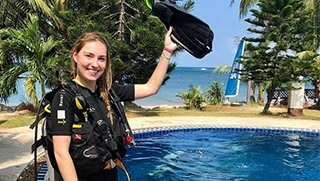 Smiling girl standing in scuba diving equipment holding one hand up with a fin in it on the side of a swimming pool close to the sea